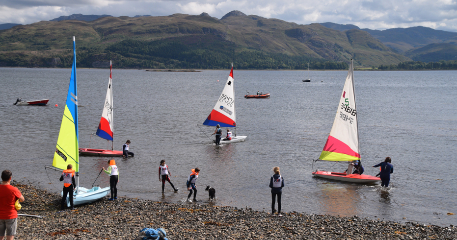 Long shot of group of sailors and their dinghys at a stone beach
